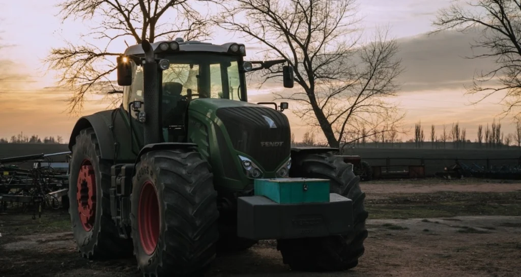 Трактор Fendt на фоні заходу сонця, стоїть на фермі серед дерев.