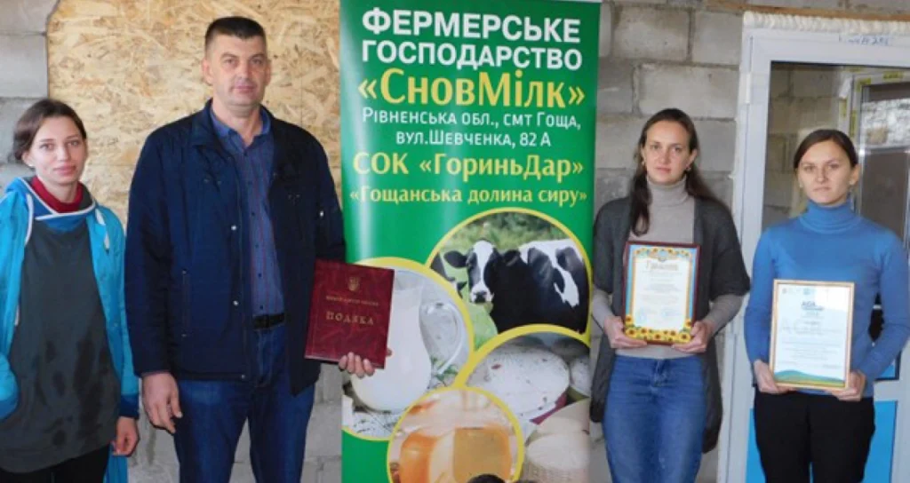 Чотири людини стоять перед банером фермерського господарства «СновМілк», тримаючи сертифікати та подяки.